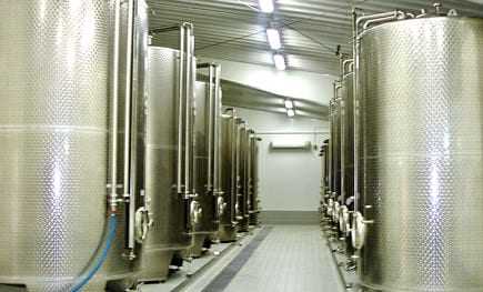 Row of stainless steel storage tanks in an industrial facility with pipes and valves. - Olive Oil Times