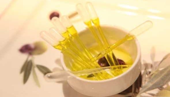 A small white bowl containing yellow oil and several clear pipettes arranged inside it. - Olive Oil Times