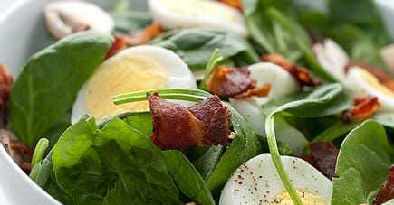 A bowl of salad containing spinach, sliced hard-boiled eggs, and pieces of bacon. - Olive Oil Times