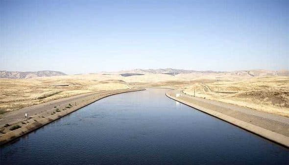 Irrigation canal running through a dry landscape in California with mountains in the background. - Olive Oil Times