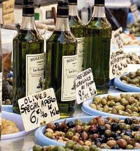 Several bottles of olive oil displayed at a market stall with various types of olives in bowls. - Olive Oil Times