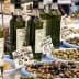Several bottles of olive oil displayed at a market stall with various types of olives in bowls. - Olive Oil Times