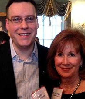 Two individuals smiling and posing for a photo at an indoor event with a decorative background. - Olive Oil Times