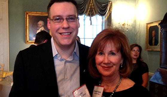 Two individuals smiling and posing for a photo at an indoor event with a decorative background. - Olive Oil Times
