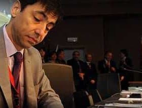 A man in a suit signing documents at a conference table with a serious expression. - Olive Oil Times