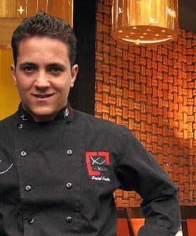 Chef wearing a black culinary uniform with a logo, standing in a restaurant setting. - Olive Oil Times