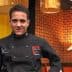 Chef wearing a black culinary uniform with a logo, standing in a restaurant setting. - Olive Oil Times
