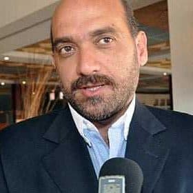 Man wearing a suit speaking into a microphone in an indoor setting. - Olive Oil Times