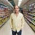 A man in a striped shirt standing in the aisle of a grocery store surrounded by various packaged products. - Olive Oil Times