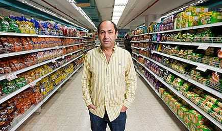 A man in a striped shirt standing in the aisle of a grocery store surrounded by various packaged products. - Olive Oil Times