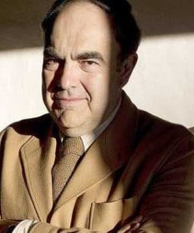 A man wearing a brown suit with arms crossed, sitting in a well-lit area with shadows. - Olive Oil Times