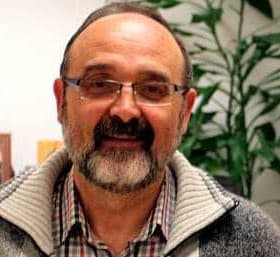 A man with glasses and a beard wearing a gray sweater, smiling in an indoor setting. - Olive Oil Times