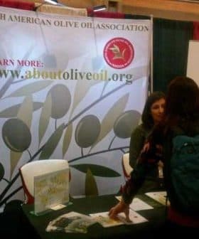 A booth for the North American Olive Oil Association with a banner and a person interacting with staff. - Olive Oil Times