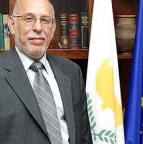 A man in a gray suit standing next to a flag of Cyprus and the European Union flag. - Olive Oil Times