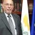 A man in a gray suit standing next to a flag of Cyprus and the European Union flag. - Olive Oil Times