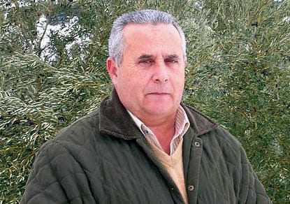 A man wearing a green jacket and beige sweater standing outdoors in front of greenery. - Olive Oil Times