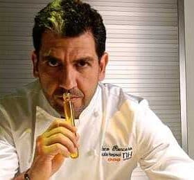 Chef holding a culinary tool while looking directly at the camera in a professional kitchen setting. - Olive Oil Times