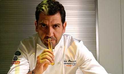 Chef holding a culinary tool while looking directly at the camera in a professional kitchen setting. - Olive Oil Times