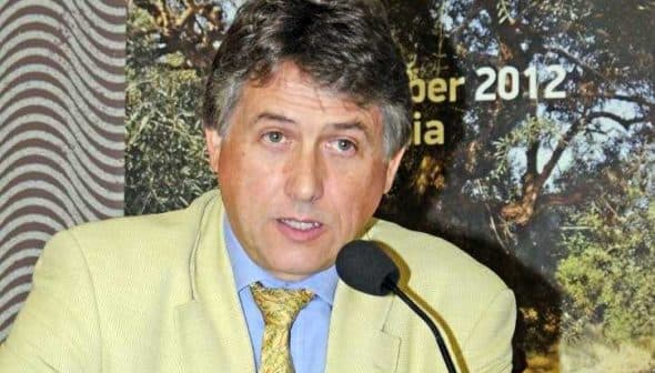 A man in a light-colored suit speaking into a microphone at a conference event. - Olive Oil Times