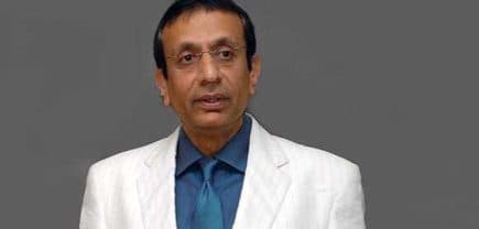 A man wearing a white suit with a blue shirt and tie, standing against a gray background. - Olive Oil Times