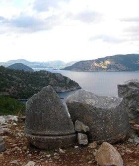 Three large stone fragments positioned near the coastline with a view of the sea and mountains in the background. - Olive Oil Times
