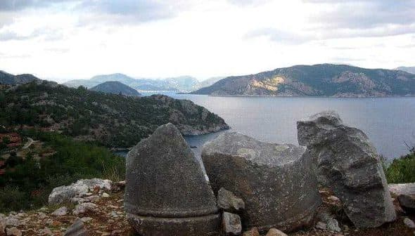 Three large stone fragments positioned near the coastline with a view of the sea and mountains in the background. - Olive Oil Times