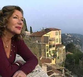 A woman with curly hair wearing a maroon top, looking out over a coastal landscape with buildings in the background. - Olive Oil Times