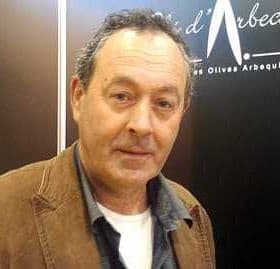 Man with dark hair and a light shirt, wearing a brown jacket, standing in front of a dark background. - Olive Oil Times