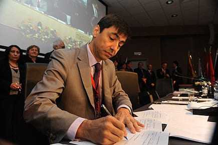 A man in a suit signing documents at a conference table with others in the background. - Olive Oil Times