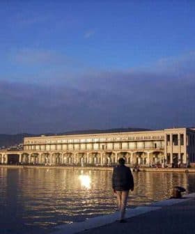 Trieste Maritime Station building reflecting on the water during sunset with a person walking nearby. - Olive Oil Times