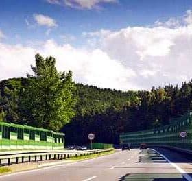 A highway with a green sound barrier on one side and trees in the background under a partly cloudy sky. - Olive Oil Times