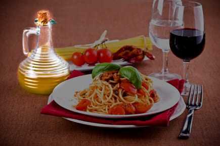 A plate of spaghetti topped with tomatoes and basil, accompanied by a cruet of olive oil and glasses of water and red wine. - Olive Oil Times