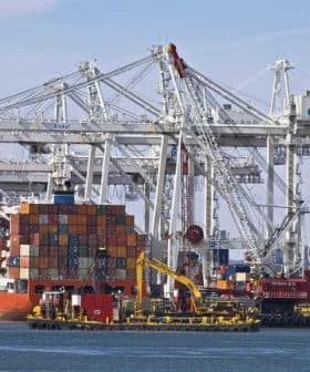 Cargo ship docked at a port with stacked shipping containers and cranes in the background. - Olive Oil Times
