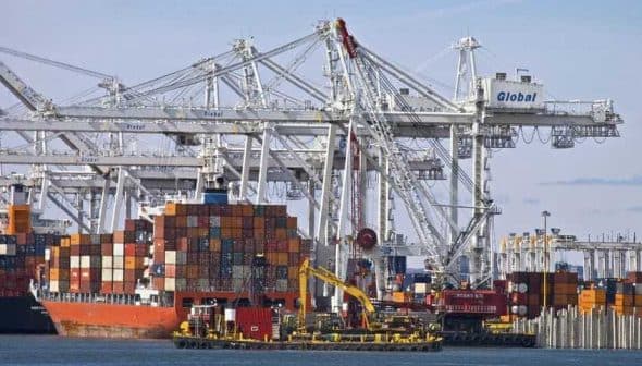 Cargo ship docked at a port with stacked shipping containers and cranes in the background. - Olive Oil Times
