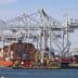 Cargo ship docked at a port with stacked shipping containers and cranes in the background. - Olive Oil Times