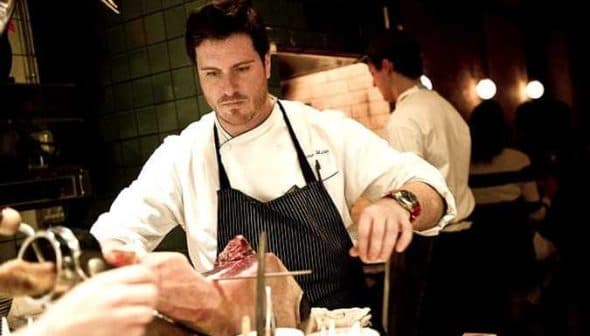 Chef in a white coat and apron preparing a piece of meat in a kitchen setting. - Olive Oil Times