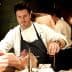 Chef in a white coat and apron preparing a piece of meat in a kitchen setting. - Olive Oil Times