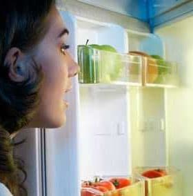 A person with dark hair looking into an open refrigerator filled with various food items and a bottle of oil. - Olive Oil Times