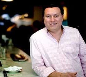 A man wearing a checkered shirt stands at a bar with a neutral expression. - Olive Oil Times