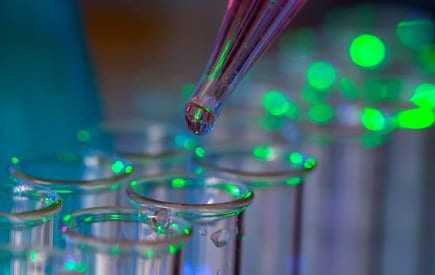 A pipette dropping liquid into a row of clear test tubes illuminated with green light. - Olive Oil Times