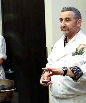 Chef in a white coat demonstrating cooking techniques with a pot in front of him and a female chef in the background. - Olive Oil Times