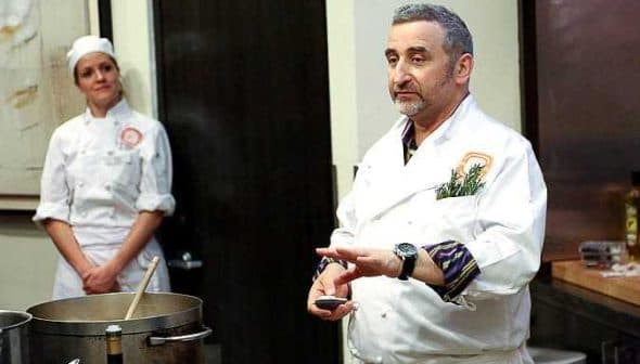 Chef in a white coat demonstrating cooking techniques with a pot in front of him and a female chef in the background. - Olive Oil Times