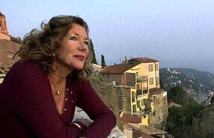 Woman with curly hair wearing a maroon top, leaning on a wall with a coastal village in the background. - Olive Oil Times
