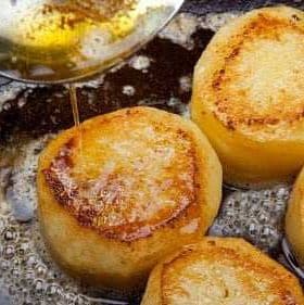 Golden-brown fried potato slices being drizzled with oil in a frying pan. - Olive Oil Times
