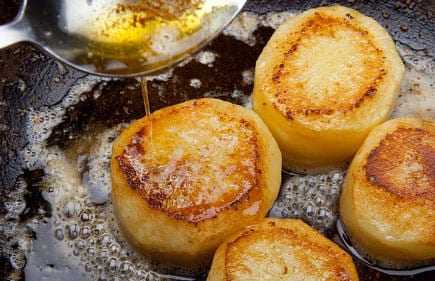 Golden-brown fried potato slices being drizzled with oil in a frying pan. - Olive Oil Times