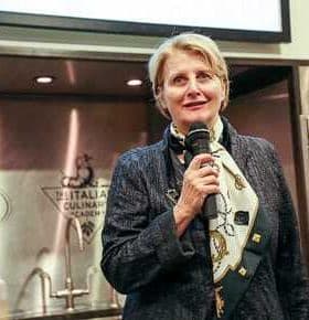 Woman holding a microphone and speaking at a culinary event with a scarf around her neck. - Olive Oil Times
