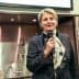 Woman holding a microphone and speaking at a culinary event with a scarf around her neck. - Olive Oil Times