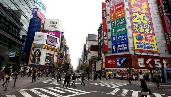 A busy intersection in Tokyo featuring large advertisements and pedestrians crossing the street. - Olive Oil Times