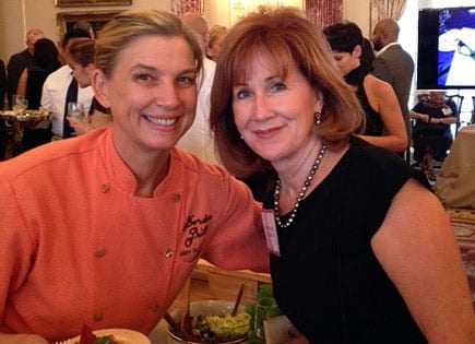 Two women smiling and posing for a photo at a social event, one wearing an orange chef's jacket. - Olive Oil Times