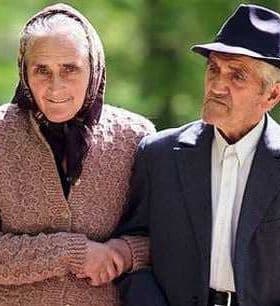 Elderly couple holding hands while walking outdoors, dressed in traditional clothing. - Olive Oil Times